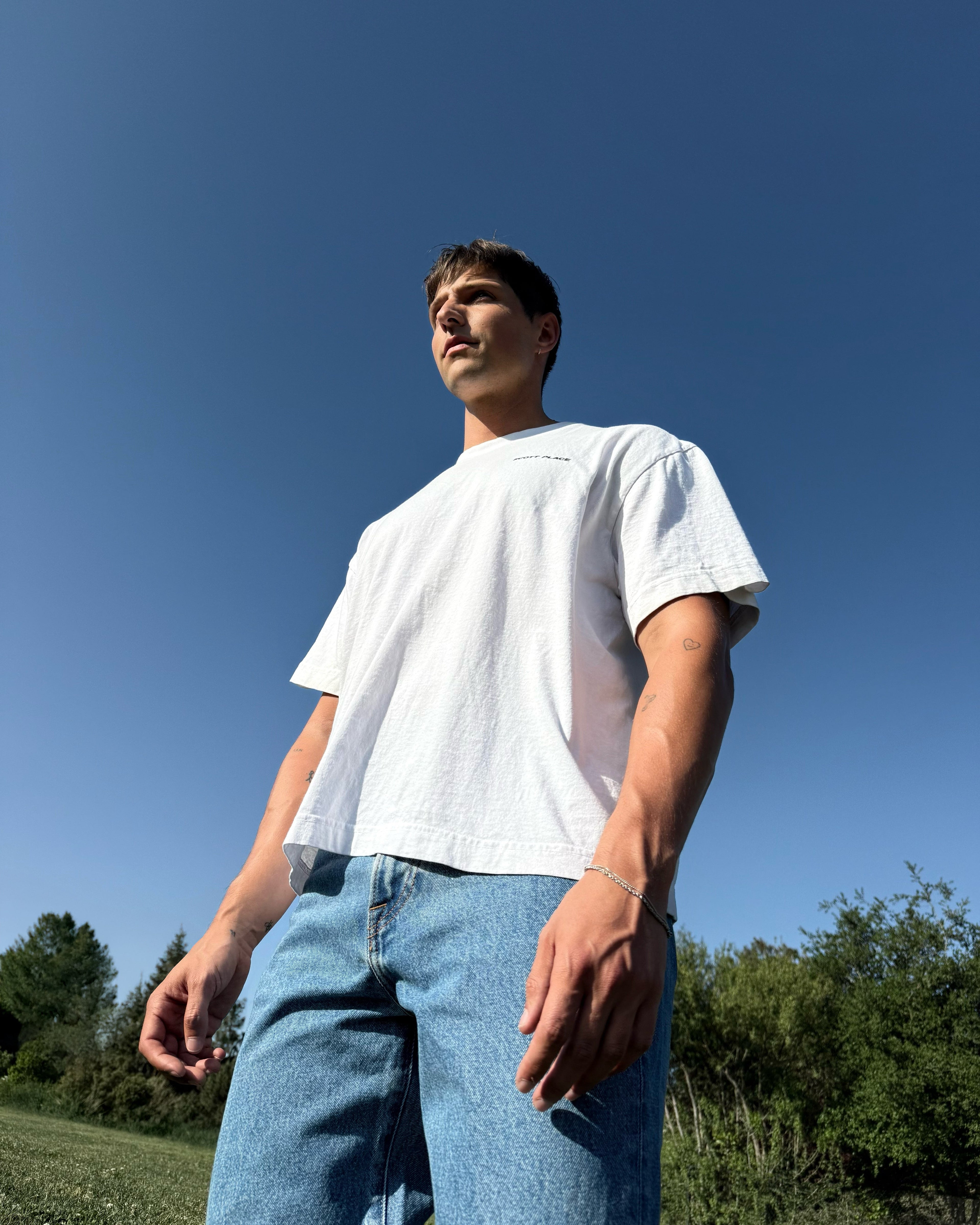 Person wearing a white t-shirt and blue jeans standing against a clear blue sky.